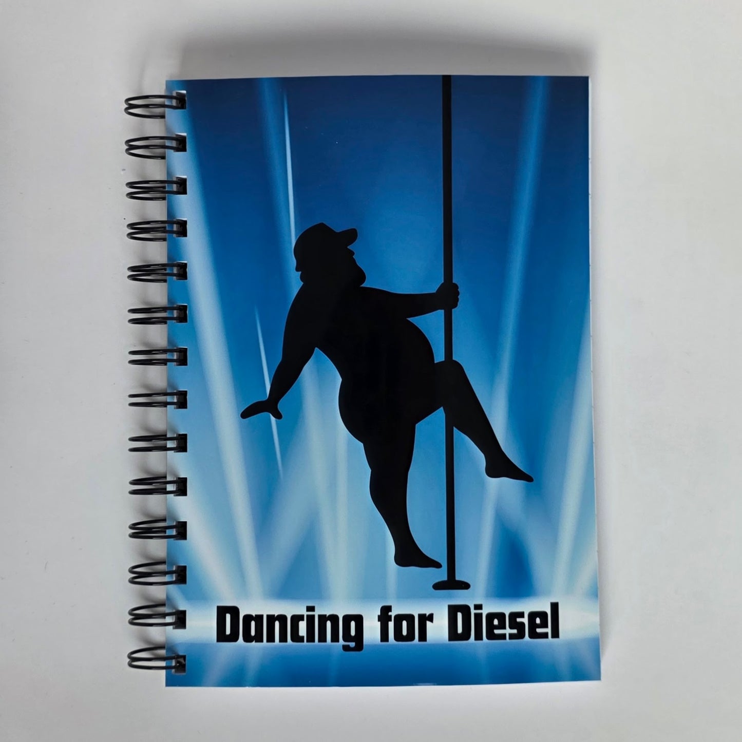 Funny notebook cover with silhouette of plus-sized cowboy pole dancing and the phrase “Dancing for Diesel” on a blue spotlight background