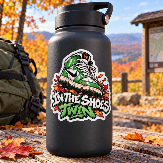 A matte black insulated water bottle with a carry loop, featuring a colorful graphic of a green and white sneaker with autumn leaves and the text 'IN THE SHOES TWIN'. The bottle is set on a wooden table outdoors with fallen leaves, a backpack, and a scenic mountain background.