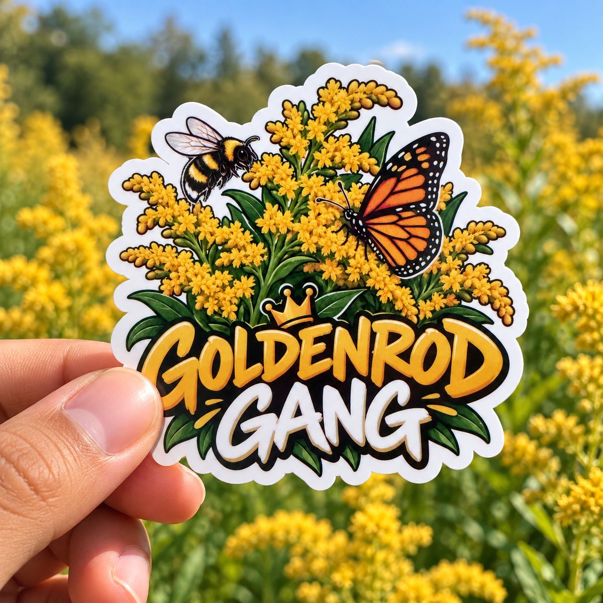 A hand holds a die-cut sticker featuring a goldenrod flower cluster, a bee, a monarch butterfly, and the words “GOLDENROD GANG” in large yellow letters, with a blurred field of yellow flowers in the background.