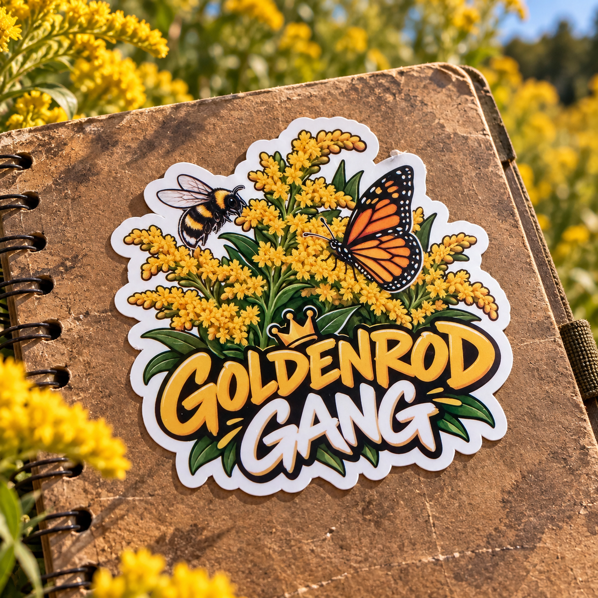 A die-cut sticker featuring a bumblebee, a monarch butterfly, yellow wildflowers, and the text 'GOLDENROD GANG' with a small crown, shown placed on the cover of a brown spiral notebook outdoors among yellow flowers.