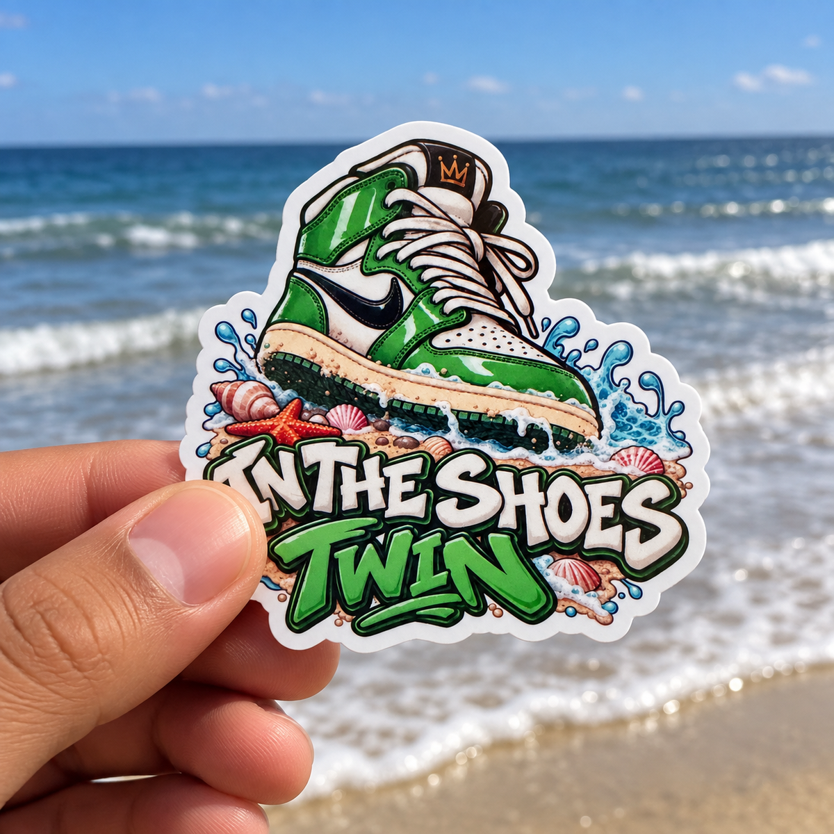 A hand holds a die-cut sticker featuring a green and white sneaker illustration with the text 'IN THE SHOES TWIN' against a blurred beach and ocean background.