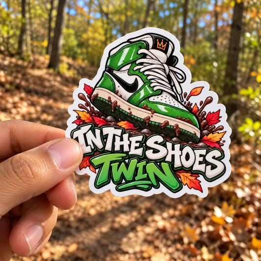 A hand holds a die-cut sticker featuring a green and white sneaker surrounded by autumn leaves, with the text 'IN THE SHOES TWIN' in bold white and green lettering. The background shows a blurred forest path in the fall.