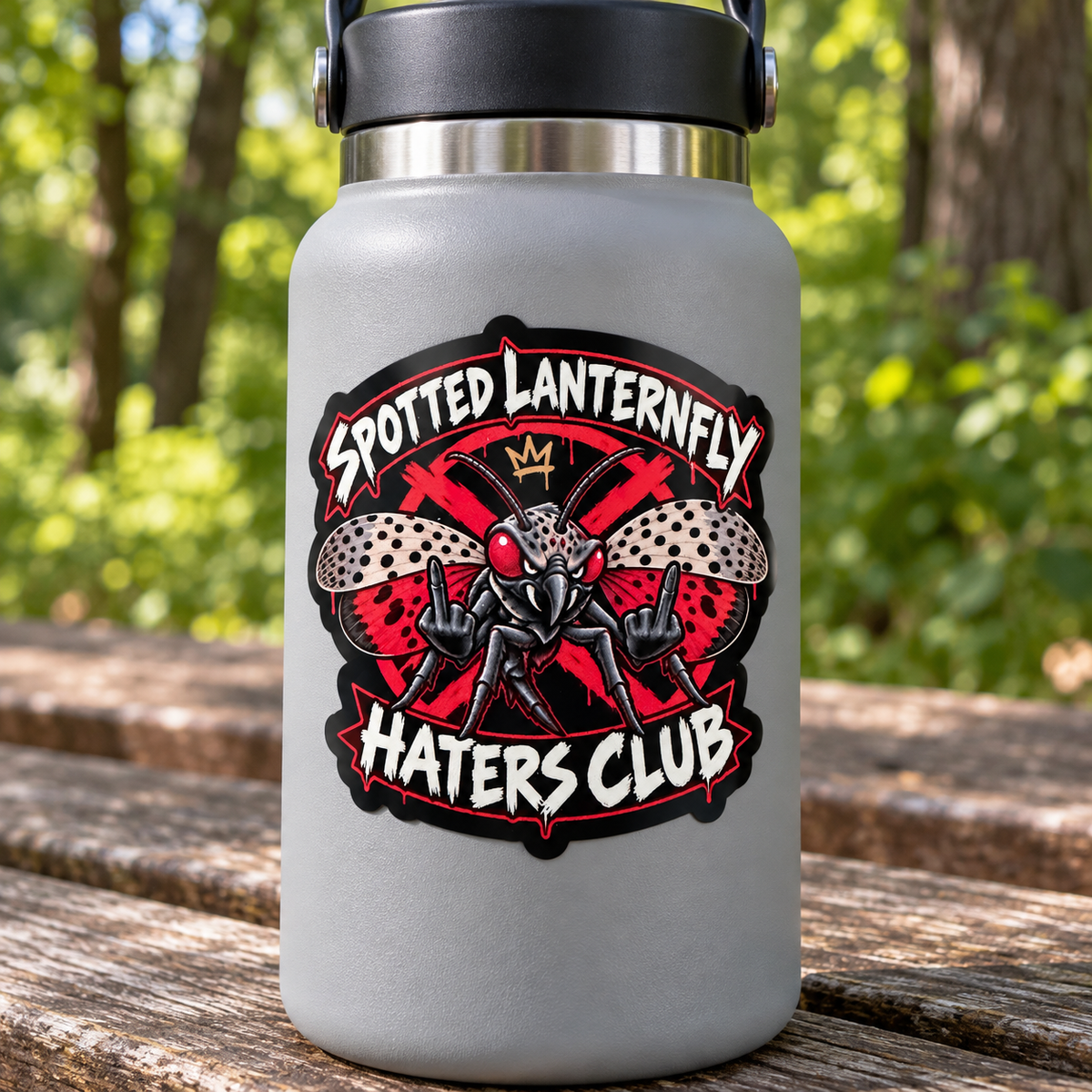 A gray insulated water bottle with a black lid sits on a wooden picnic table outdoors. The bottle features a large graphic design with the text 'Spotted Lanternfly Haters Club' and an illustration of a lanternfly with red wings.